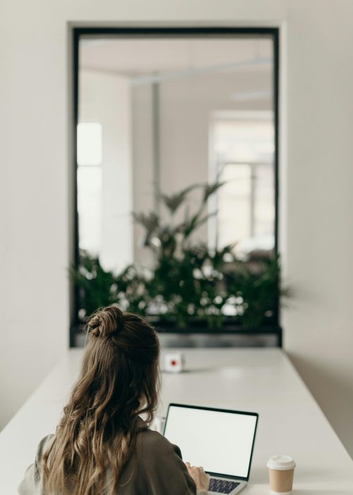 pexels-photo-4065624-4065624 A woman with curly hair works on a laptop, facing a window with plants, symbolizing remote work.